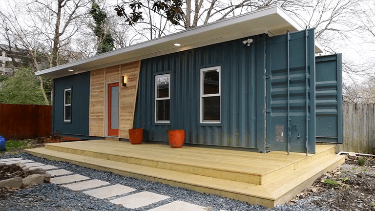 Exterior of a metal shipping container retrofited as a home with three windows and a wooden door on the facing side.